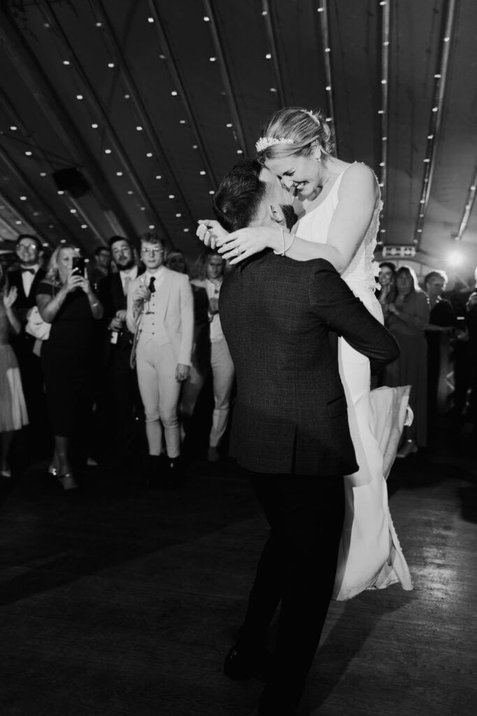 Groom lifts bride during joyful first dance at Thorpe Garden winter wedding, surrounded by cheering guests under twinkling lights.