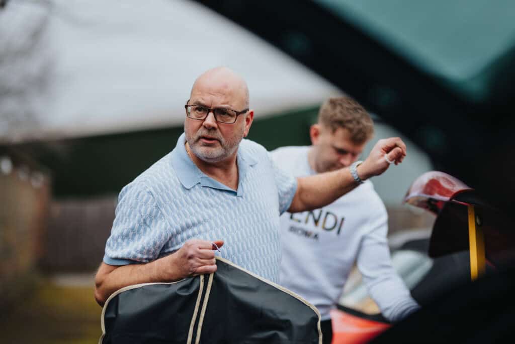 Father of the bride helps unload wedding items from the car during morning preparations at Thorpe Garden winter wedding, with a groomsman assisting in the background.