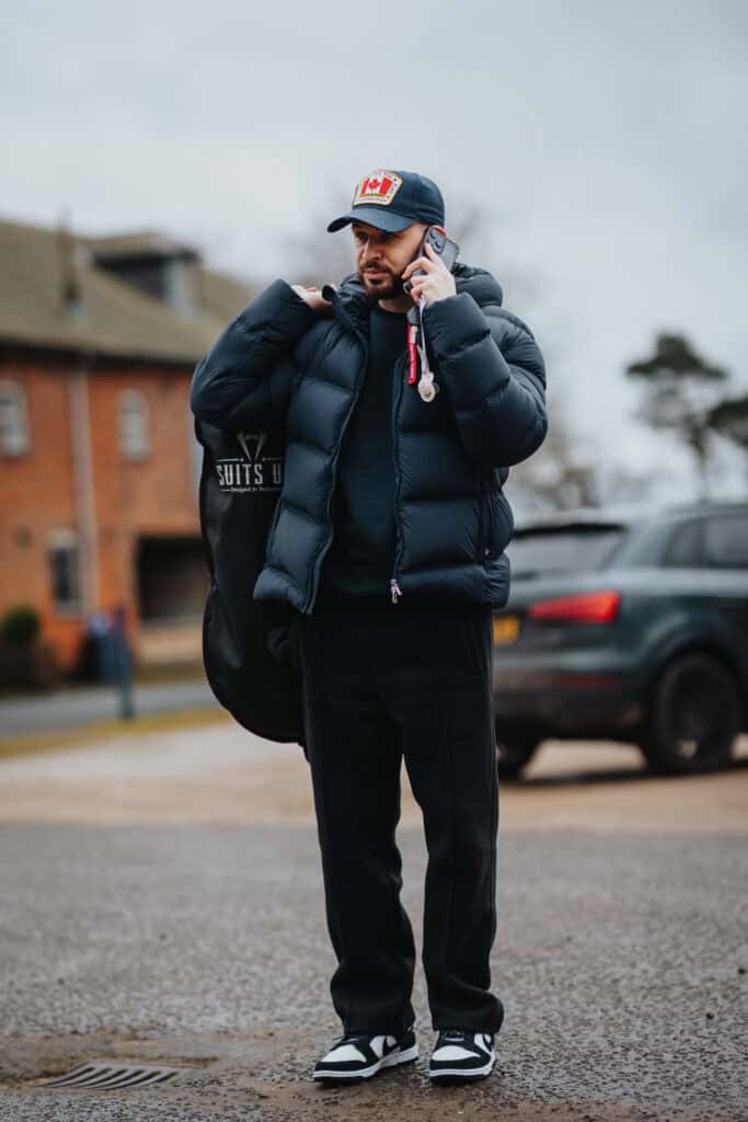 Groom talking on the phone while carrying a suit bag during wedding morning preparations at Thorpe Garden, dressed in a puffer jacket