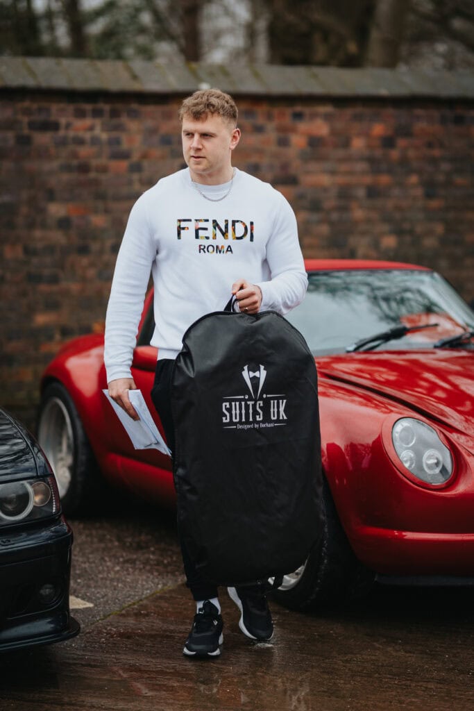 Groomsman holding a suit bag and wedding notes while arriving at Thorpe Garden for winter wedding preparations, standing by a red sports car.