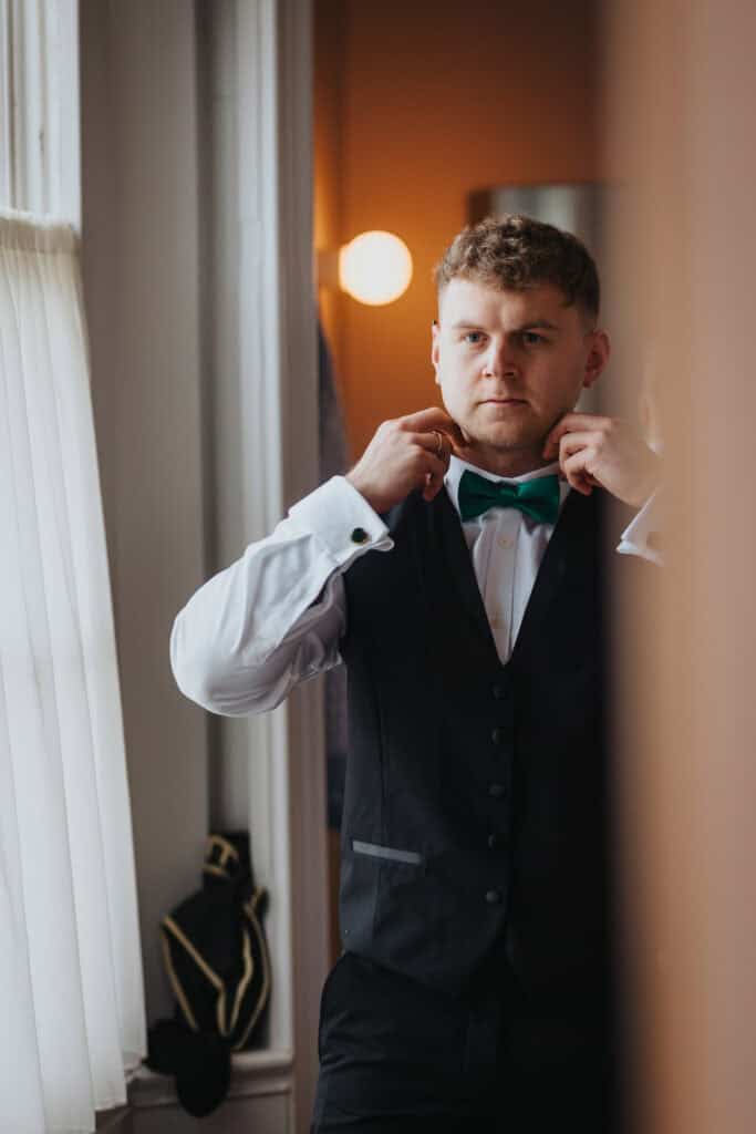 Groomsman fixing his green bow tie in front of a window while getting ready for a winter wedding at Thorpe Garden in Staffordshire.