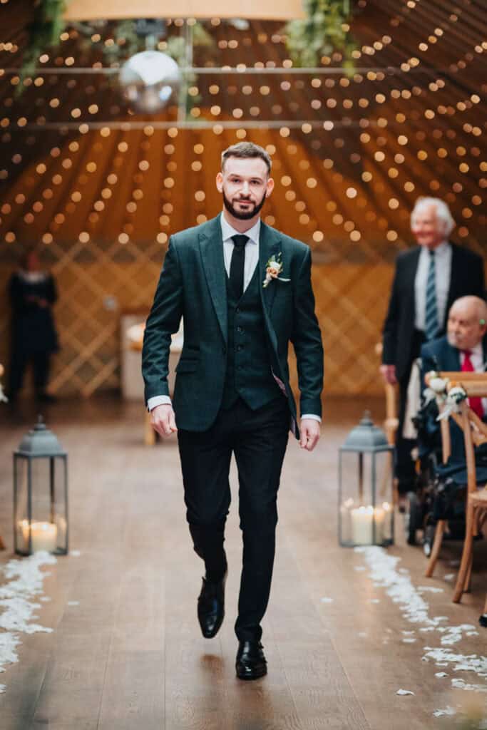 Groom walking down the aisle at Thorpe Garden wedding ceremony in Staffordshire, surrounded by warm fairy lights and rustic wooden decor during a February winter wedding.