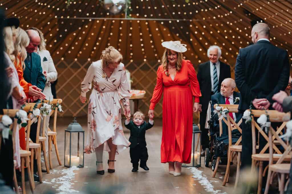 Bride and groom's young son walking down the aisle holding hands with two family members during the wedding ceremony at Thorpe Garden, Staffordshire.