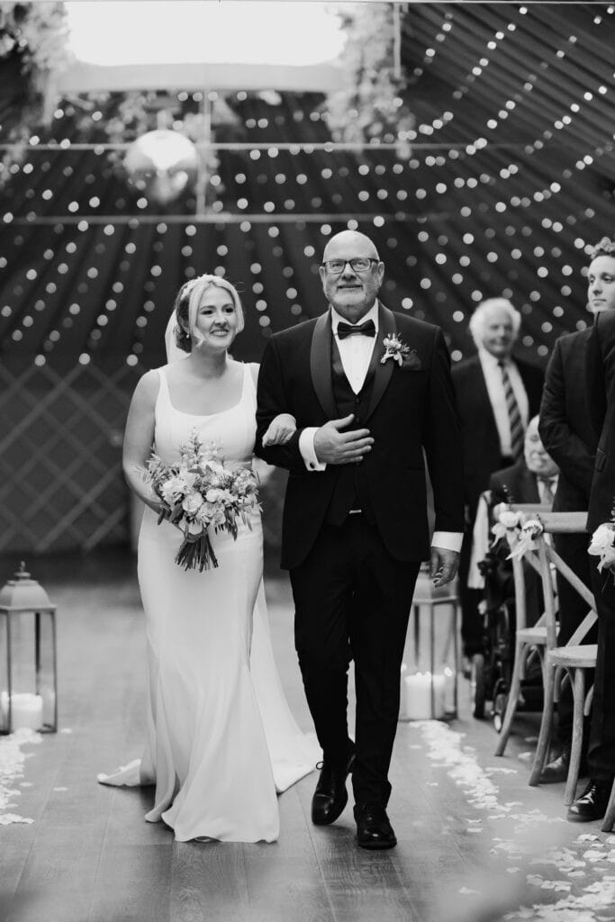 Bride walking arm in arm with her father down the aisle during a winter wedding ceremony at Thorpe Garden, Staffordshire, with soft fairy lights in the background.