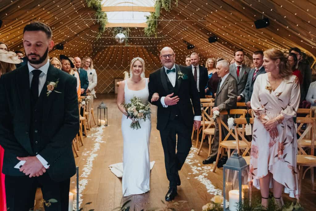 Bride walks down the aisle with her father at Thorpe Garden winter wedding, holding a bouquet, surrounded by smiling guests and fairy lights.