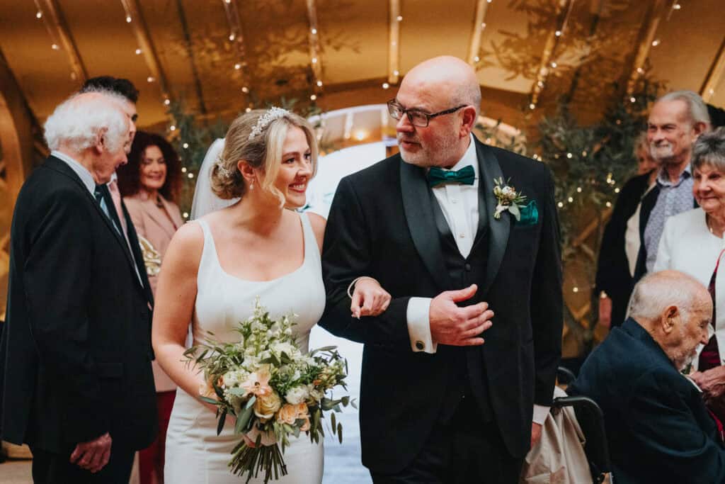 Bride and her father share a smile while walking down the aisle at Thorpe Garden winter wedding, holding a fresh bouquet surrounded by guests.