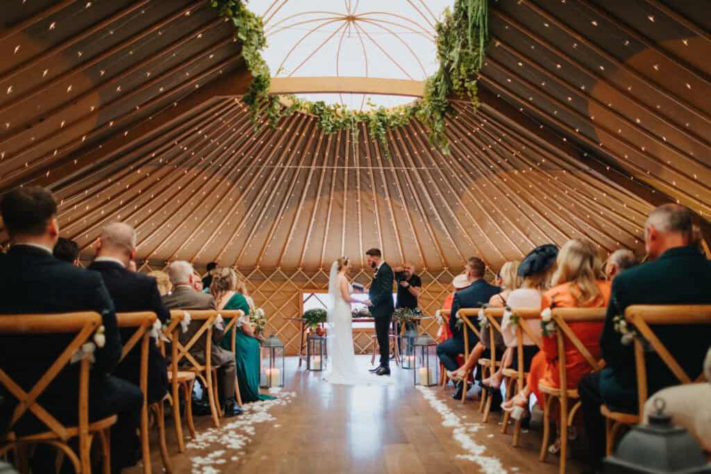 Bride and groom exchanging vows at Thorpe Garden ceremony with guests seated and wedding photographer Marcin smiling in the background.