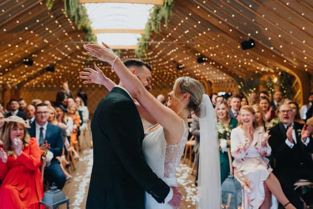 Bride and groom sharing their first kiss during the wedding ceremony at Thorpe Garden, Staffordshire, surrounded by cheering family and friends.