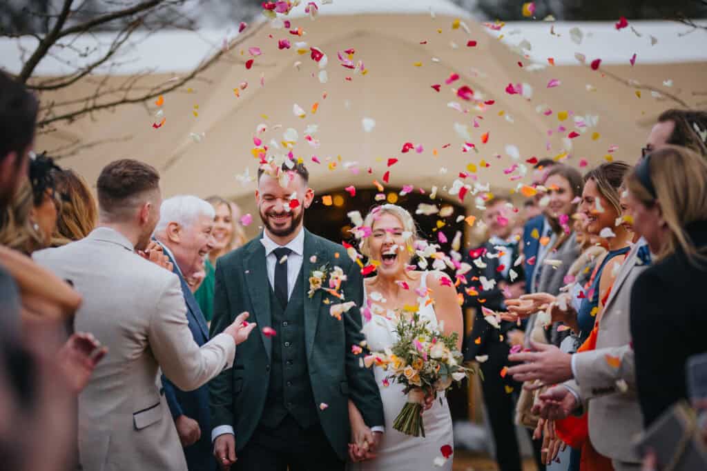 Bride and groom walk hand in hand through colourful confetti shower during outdoor winter wedding at Thorpe Garden, surrounded by cheering guests.