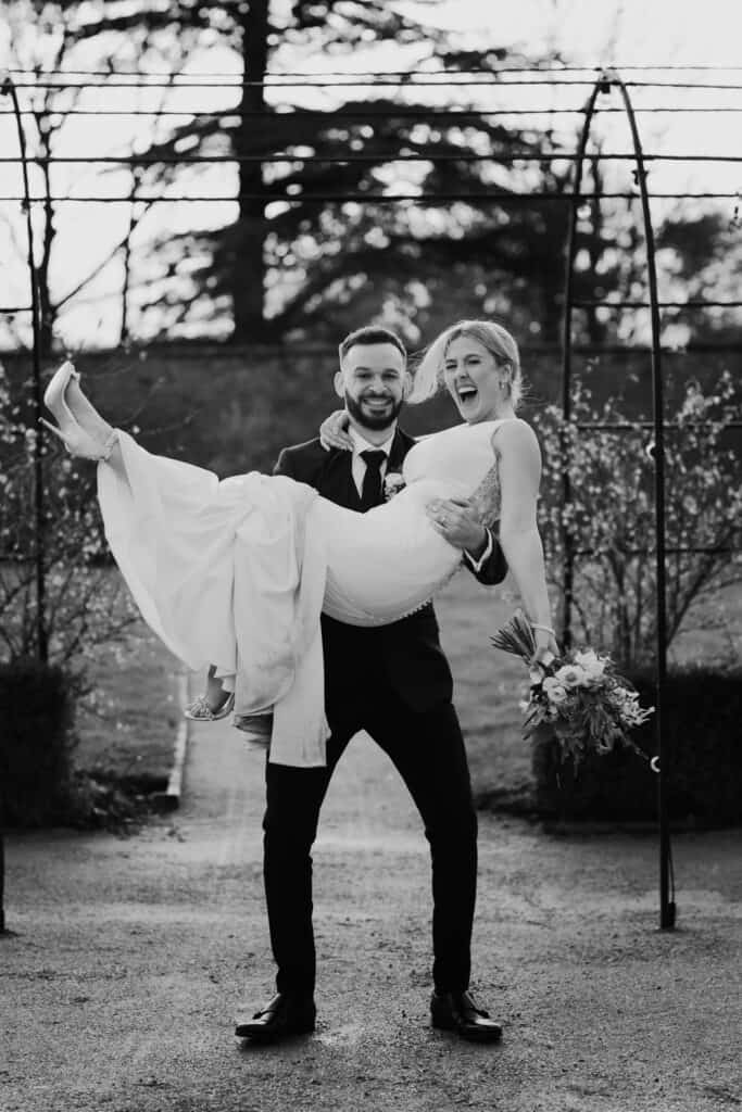 Groom lifts smiling bride holding bouquet during playful wedding portrait at Thorpe Garden in winter, with trees and garden arches in the background.