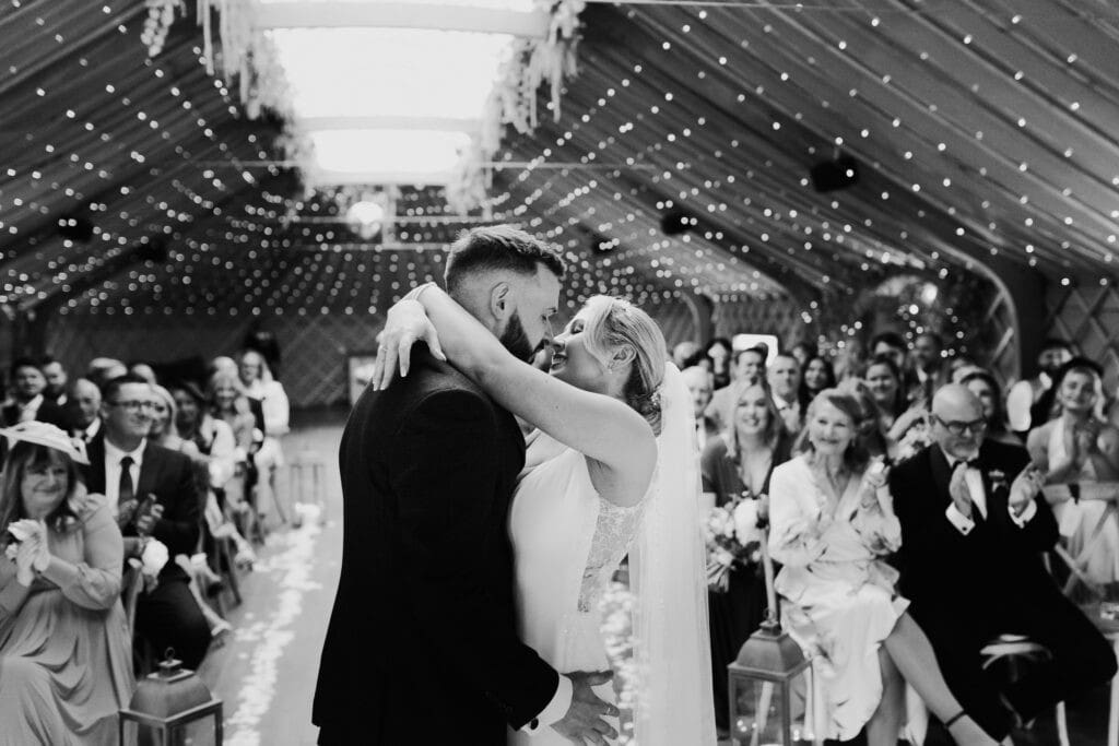 Bride and groom share emotional first kiss under fairy lights at Thorpe Garden winter wedding, surrounded by smiling guests.