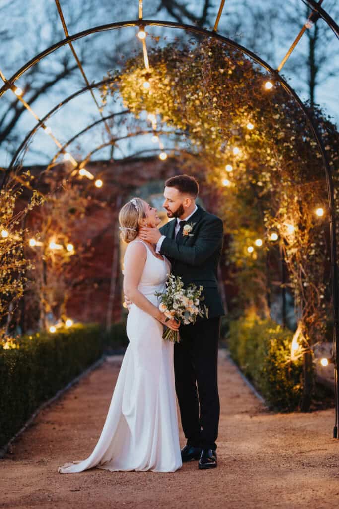 Bride and groom share a romantic moment under glowing archway lights during their winter wedding at Thorpe Garden, with soft evening light and greenery.