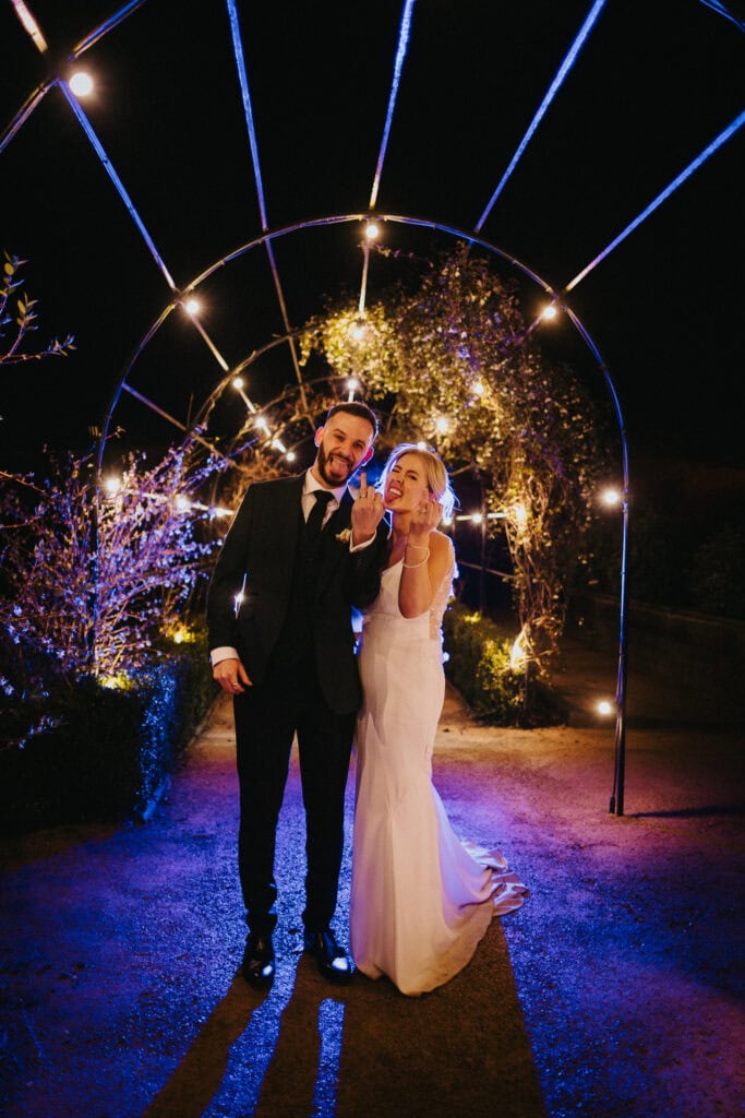 Bride and groom at Thorpe Garden posing under evening lights, sharing a cheeky moment during their wedding day with playful expressions.