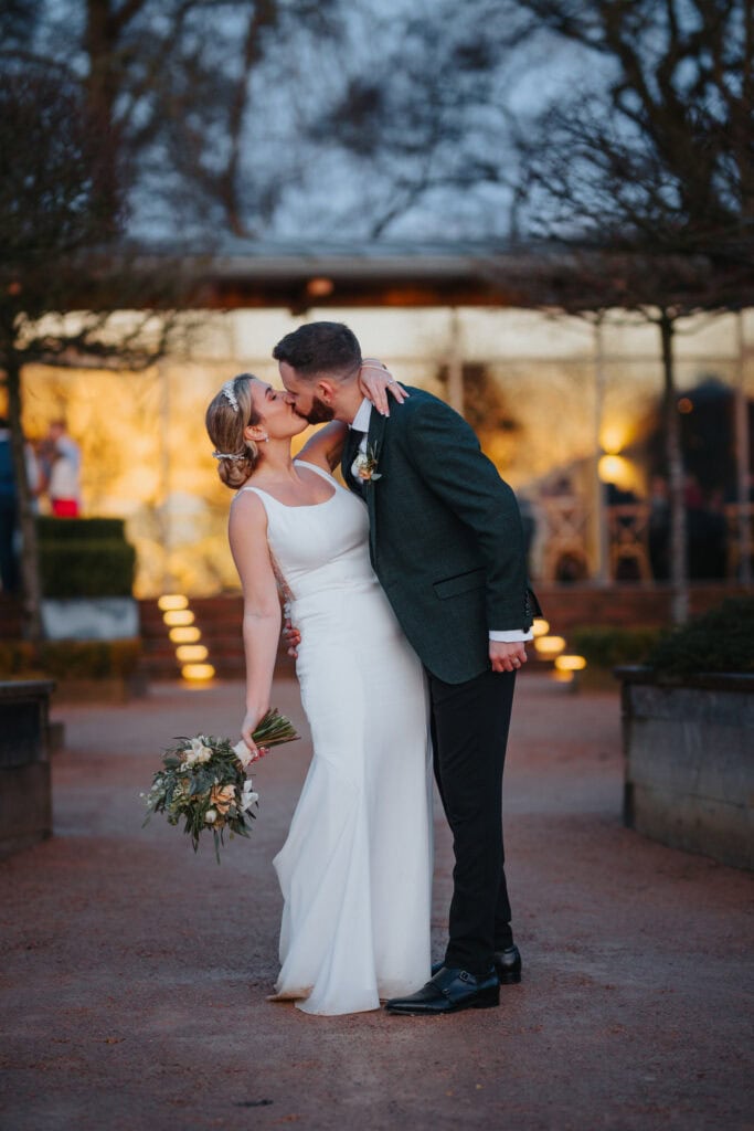 Bride and groom share a romantic kiss at sunset during their winter wedding at Thorpe Garden, with soft golden light and garden details in the background.