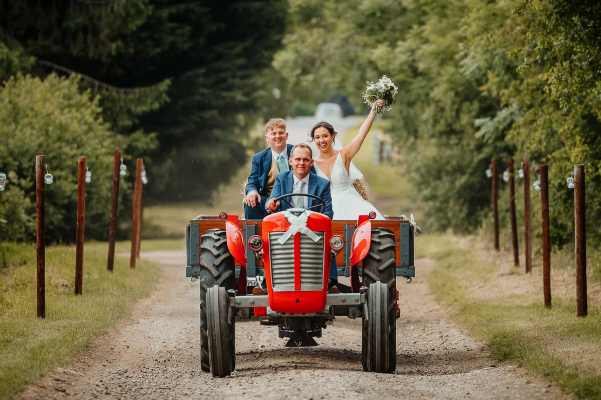 Bride and groom arriving on a red tractor with a guest, holding bouquet in the air
